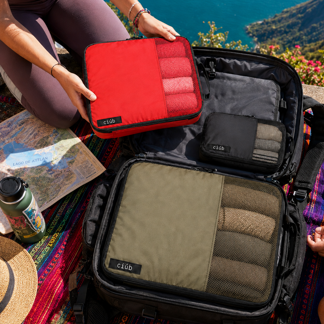 A travel organizer set from the brand Club is shown packed into and around an open suitcase on a colorful picnic blanket with a scenic mountain and ocean background. The set includes a red zippered case with folded towels, a black zippered case with folded towels, and a large olive green organizer pouch with mesh pockets holding brown and beige towels.